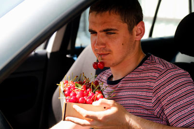Fun caucasian young man sitting in car and holding sweet cherry in box. 20s guy driver looking at 