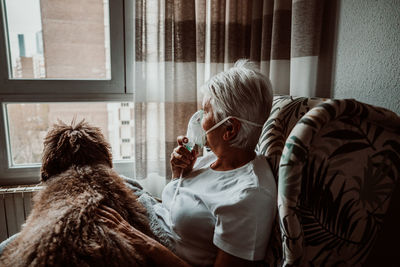 Senior woman wearing oxygen mask sitting with dog on chair at home