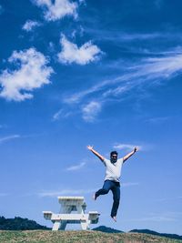 Low angle view of man with arms raised jumping from picnic table against blue sky