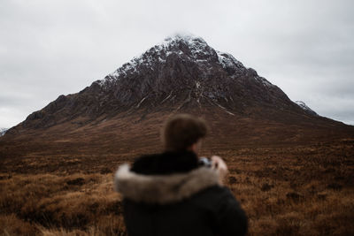Back view of unrecognizable blurred tourist in warm clothes admiring landscape of mountain with snowy peak in scottish highlands in fall