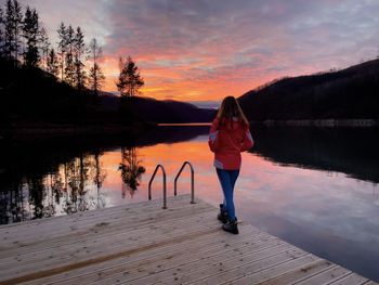 Woman standing on a wooden deck over the lake and watching an amazing sunset reflecting in the water