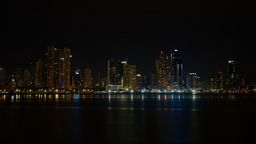 Illuminated buildings by river against sky at night