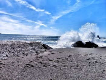 Scenic view from beach and waves crashing on boulders against sky