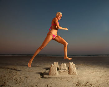 Happy mid adult woman jumping over sandcastle at beach during dusk