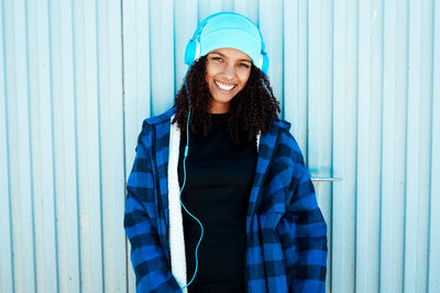 Portrait of smiling young woman listening to music on headphones while standing against closed shutter