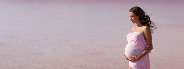 Pregnant woman stands on sea beach in summer in white light dress with fluffy