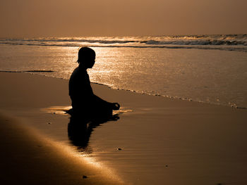 Silhouette boy meditating while sitting on sand at beach against sky during sunset