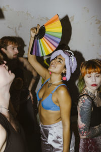 Smiling non-binary person holding multicolored hand fan and dancing with friends at nightclub