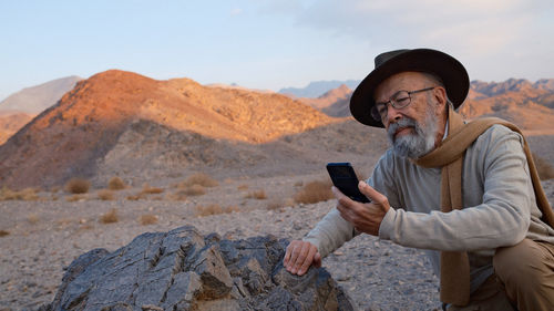 Young man using mobile phone while standing on rock against sky