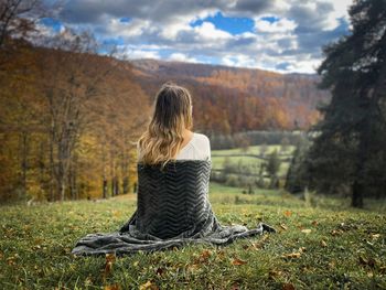 Woman covered with a blanket sitting down on the grass and looking at the autumn scenery around