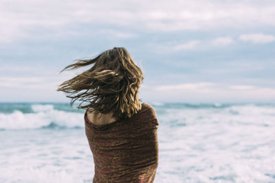 Rear view of mid adult woman with scarf standing at beach against cloudy sky