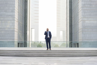 Full length of businessman talking on mobile phone while standing against office buildings