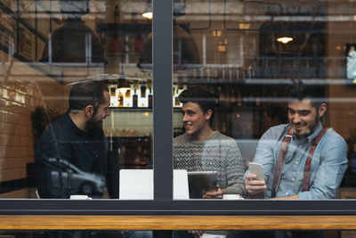 Friends working on laptop while sitting in cafe seen through window