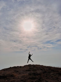 Silhouette person with arms raised jumping on hill against sky during sunset