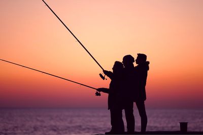 Silhouette friends fishing at beach against sky during sunset