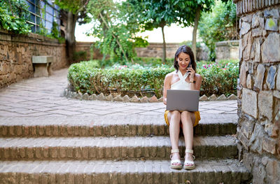 Attentive self employed woman talking phone and working on project while sitting with laptop on stairs