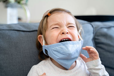 Close-up of crying toddler with mask sitting on sofa at home