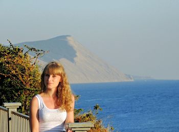 Portrait of woman standing on bridge by sea and mountain against clear sky