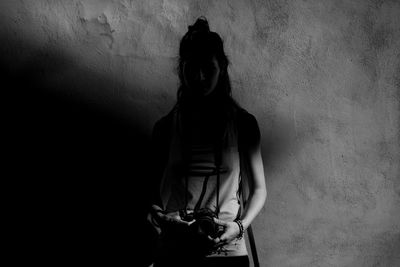 Young woman holding camera while standing against wall in darkroom