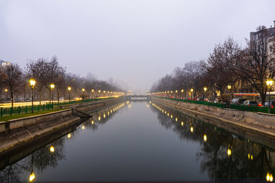 Illuminated street by lake against sky at dusk