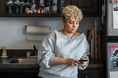 Woman with short hair text messaging on smart phone while leaning on kitchen counter at home