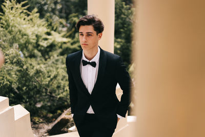 A beautiful young man, the groom in an elegant wedding suit, stands posing in the city's old park