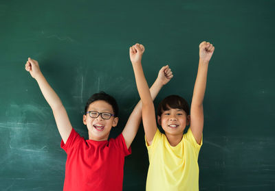 Portrait of happy friends with arms raised standing by blackboard in classroom