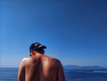 Rear view of shirtless man against sea against clear blue sky