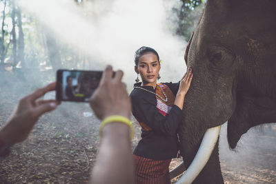 Cropped hand of person photographing woman with elephant in forest