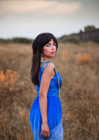 Portrait of young beautiful woman wearing blue dress on field against sky