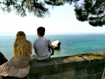 Rear view of boy and girl sitting on retaining wall against sea