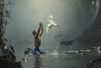 Shirtless boy with arms raised looking at bird flying while sitting on rock in river