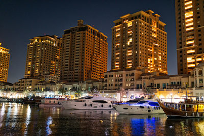 Boats moored in river by illuminated modern buildings at night