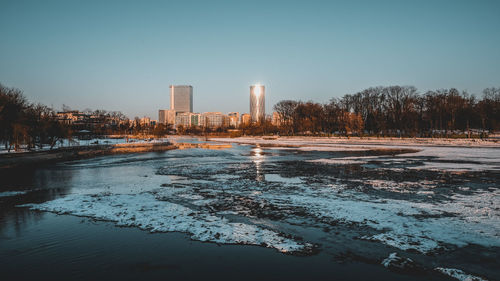 Frozen river by buildings against clear sky during winter