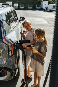Smiling siblings holding ice creams and charging car while standing at station
