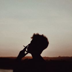 Portrait of silhouette woman holding cigarette against sky at sunset