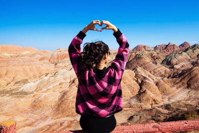 Rear view of young woman making heart shape while sitting on wooden railing against clear blue sky during sunny day