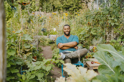 Portrait of smiling male farmer with arms crossed sitting in community garden