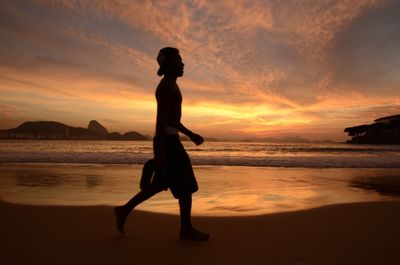 Full length of shirtless man walking at beach against sky during sunset