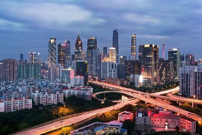 Light trails on elevated road amidst modern buildings against cloudy sky at dusk