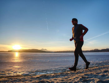 Athlete runner running on beach at frozen lake in rays of sunset