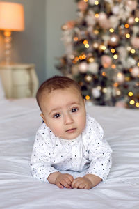 Baby boy six months lying on the bed in a white shirt next to the christmas tree