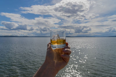 Cropped hand of person holding whisky glass against sea and cloudy sky