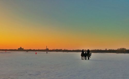 Silhouette people riding and walking with horses on snow covered field at dusk