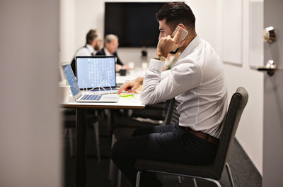 Young businessman talking on phone while using laptop at conference table in board room