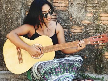 Young woman playing guitar while sitting by old wall
