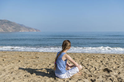 Rear view of girl sitting on beach against sky