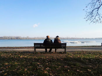 Rear view of men sitting on bench by lake against sky