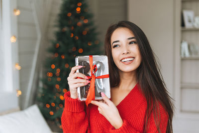 Young asian woman in red sweater and santa hat with gift box in room with christmas tree at home