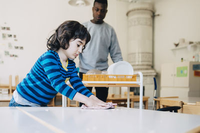 Boy cleaning table with dish cloth while teacher standing in classroom at kindergarten